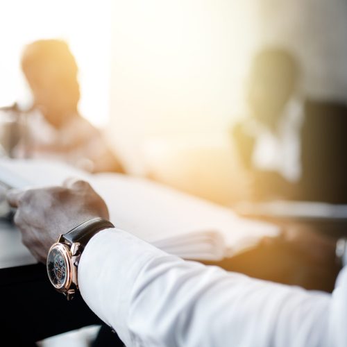 Close-up hand of a businessman in a white shirt with a clock holding documents. In the background are blurry colleagues and bright sunlight.